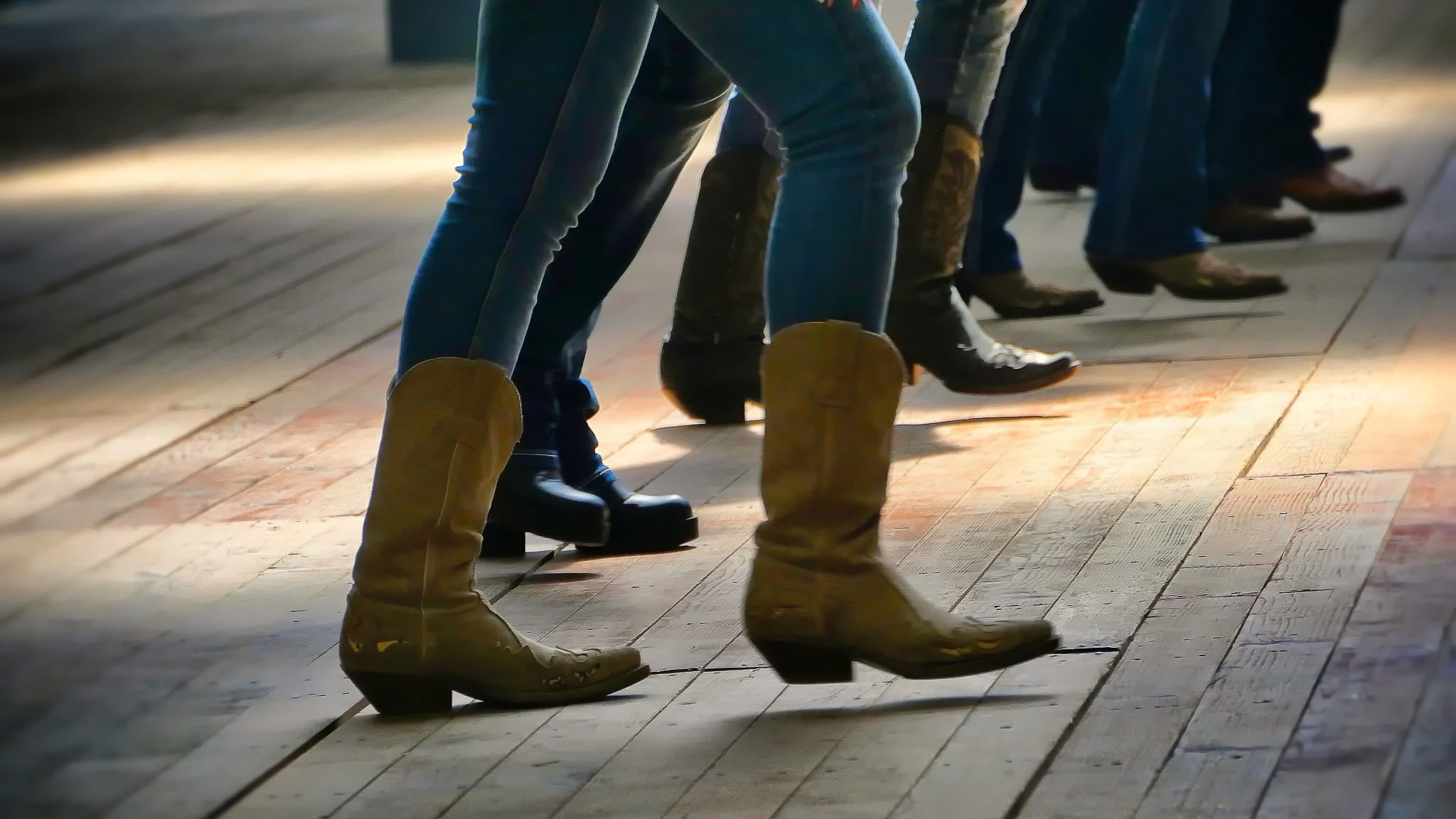 Personer i cowboyboots dansar linedance i grupp på ett trägolv.
